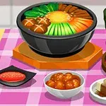 Mâncăruri diverse pe o masă cu față de masă roz, inclusiv o supă cu legume într-un bol negru, un pahar cu băutură verde, și un platou cu kimchi
