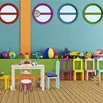 Sala de joacă colorată cu jucării, mese și scaune viu colorate, geamuri rotunde în perete și un șevalet