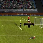 Un jucător îmbrăcat în galben execută un șut de la distanță spre o poartă pe terenul de fotbal, iar un alt jucător cade lângă poartă