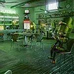Interiorul unui bar futurist unde mai mulți roboți sunt așezați la mese, înconjurați de decoruri futuriste și un semn luminos Le Robobar