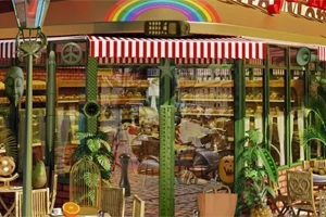 Intrare într-o cafenea cu geamuri mari, acoperiș cu dungi roșu și alb, decorată cu plante și mobilier de exterior sub un curcubeu