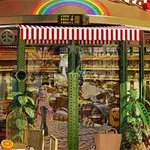 Intrare într-o cafenea cu geamuri mari, acoperiș cu dungi roșu și alb, decorată cu plante și mobilier de exterior sub un curcubeu