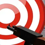 O țintă circulară roșie și albă și un pistol îndreptat către aceasta, pe un fundal de zid de piatră