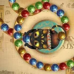 Un șir de bile colorate trece printr-un cap de broască neagră decorată cu detalii viu colorate, pe un fundal cu motive egiptene